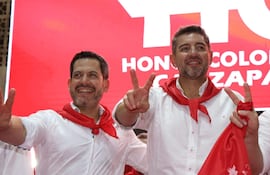 Alliana y Latorre en camisas blancas con pañuelos rojos, haciendo gestos de victoria en un fondo rojo.