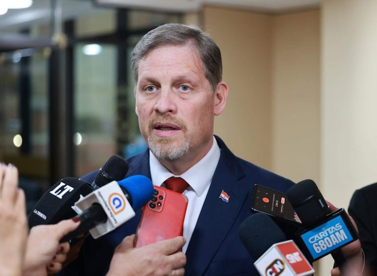 Hombre en traje oscuro y corbata roja da declaraciones a la prensa, rodeado de micrófonos y dispositivos móviles.