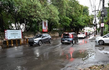 Calle Gral Santos con tráfico, charcos de agua, un automóvil gris y un SUV azul entre otros coches, bajo un cielo nublado.