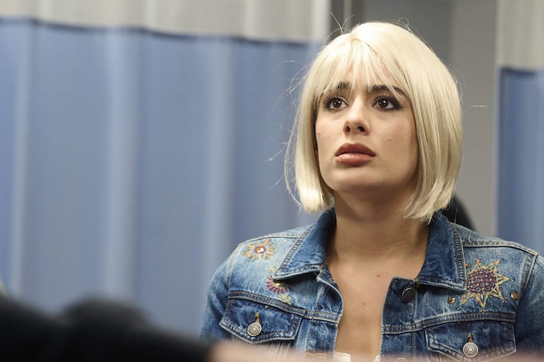 Mujer joven con cabello rubio y chaqueta de mezclilla decorada, mostrando sorpresa mientras escucha a alguien fuera de la vista.