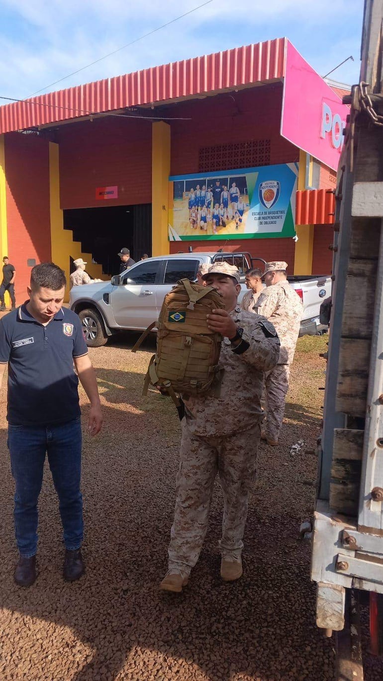 Hombre en camiseta oscura interactúa con un uniforme militar que sostiene una mochila con bandera de Brasil en una zona exterior iluminada.