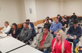 Grupo de 12 personas en sala de reunión; mujeres con atuendos formales tomando notas, expresiones de tranquilidad y tristeza.