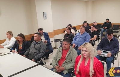 Grupo de 12 personas en sala de reunión; mujeres con atuendos formales tomando notas, expresiones de tranquilidad y tristeza.
