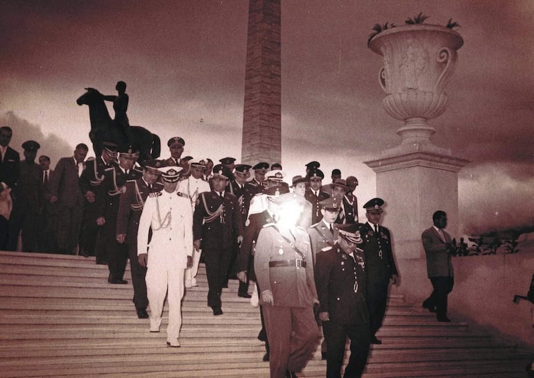 Grupo de hombres en uniformes militares y de gala, con insignias, descendiendo solemnemente por una escalera hacia un ambiente emblemático.