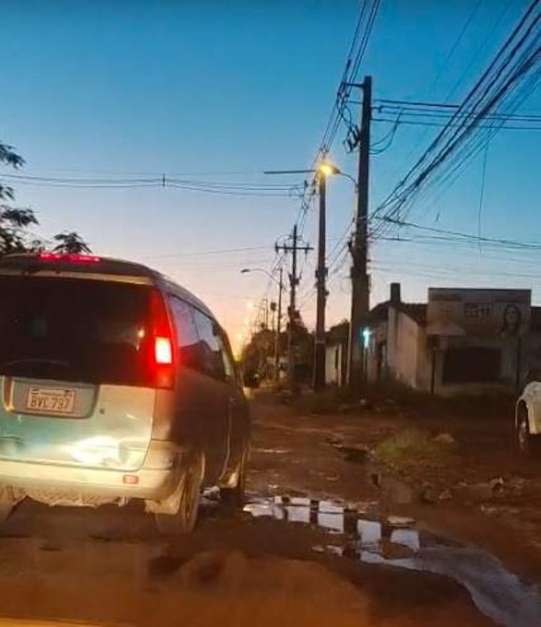Vehículo aparcado en calle con pavimento dañado y charcos, rodeado de postes de luz al anochecer.