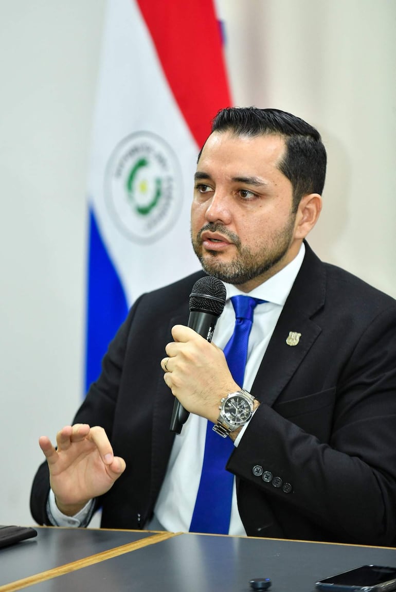 Luis Bello con traje oscuro y corbata azul sostiene un micrófono y gesticula en conferencia con bandera paraguaya de fondo.