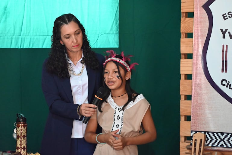 Mujer con traje formal y micrófono escucha a niña con adornos culturales y pintura facial, en un ambiente educativo interior.