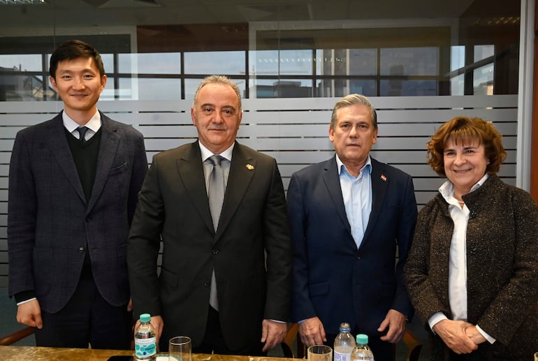 Cuatro personas alineadas en una sala: dos hombres con trajes formales, uno serio, y una mujer sonriendo con abrigo oscuro.