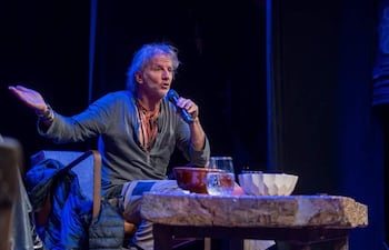 Facundo Arana con cabello claro y camiseta gris, usa un micrófono mientras gesticula en un escenario oscuro.