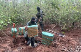 Dos personas con pasamontañas y ropa camuflada inspeccionan paquetes envueltos en plástico en un entorno forestal.