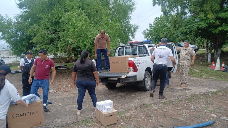 Seis personas manejan cajas con la marca 'COMITIA'; un hombre en uniforme negro observa mientras una mujer alejada lleva un paquete.