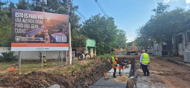Cinco trabajadores con cascos y chalecos reflectantes en obra pública, manipulando materiales y supervisando el trabajo bajo un cartel informativo.