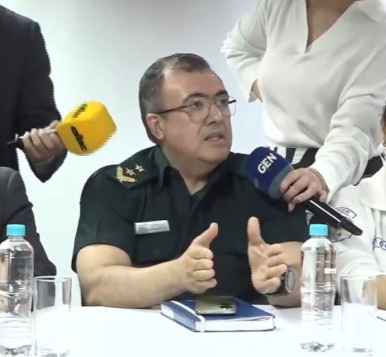 Un militar con gafas gesticulando en conferencia de prensa, rodeado de personas y micrófonos en ambiente interior.