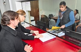Jorge Sabaté y Melissa Ramírez en oficina, revisando documentos sobre una mesa roja, en ambiente administrativo.