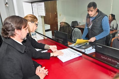 Jorge Sabaté y Melissa Ramírez en oficina, revisando documentos sobre una mesa roja, en ambiente administrativo.