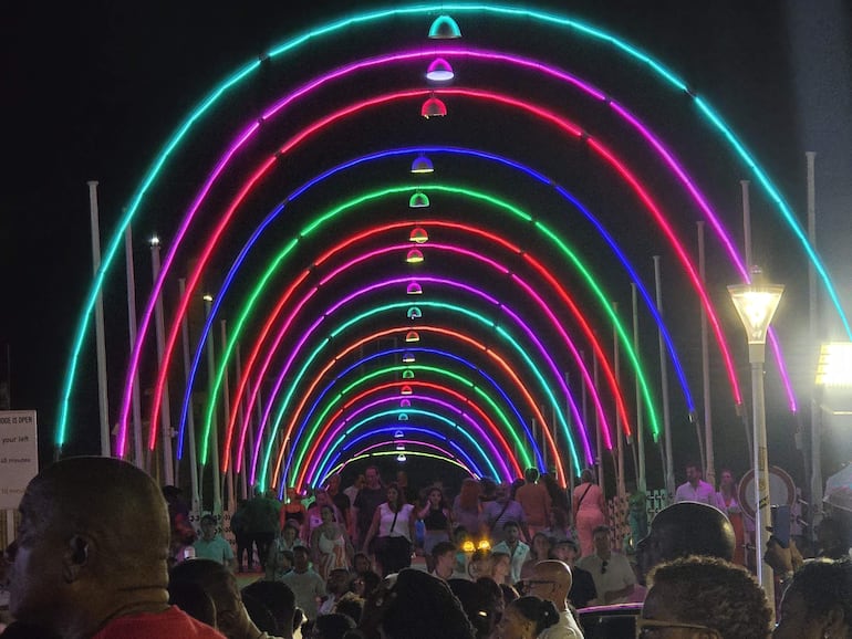 Una multitud variada bajo un arco iluminado en colores vibrantes, con personas sonriendo y disfrutando del ambiente festivo.