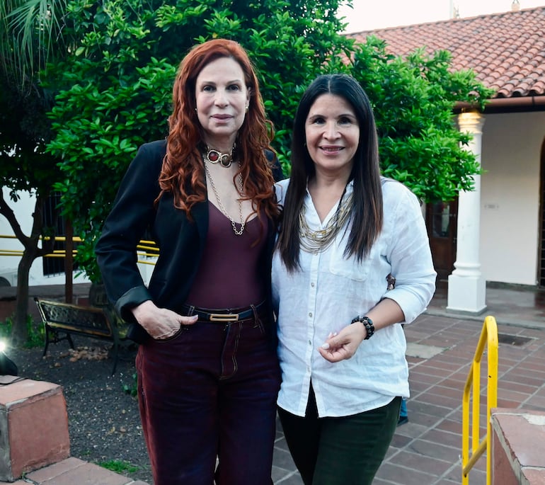 Mujeres sonriendo en un entorno al aire libre, la de izquierda con blusa oscura y pantalones burdeos, la de derecha en blusa blanca y pantalones oscuros.
