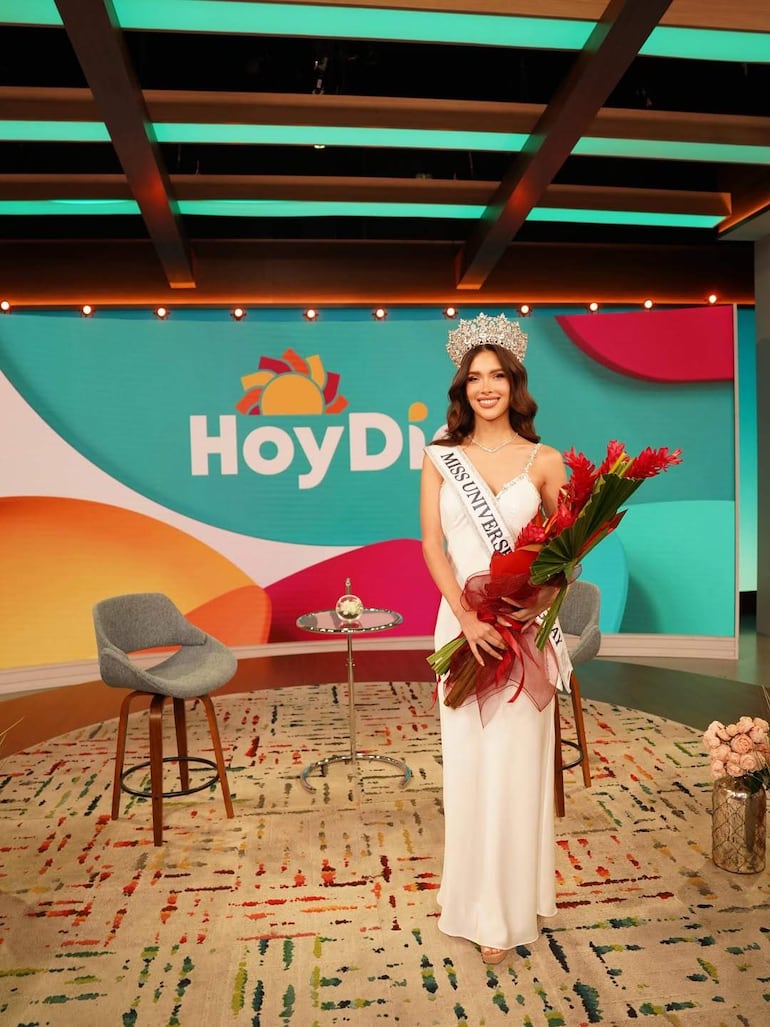 Gretha Matiauda, con tiara y vestido blanco, sostiene un ramo de flores con expresión feliz en set de televisión colorido.