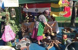 Mujer con delantal verde cocina en un fogón, mientras otros observan en un ambiente decorado con banderas paraguayas.