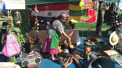 Mujer con delantal verde cocina en un fogón, mientras otros observan en un ambiente decorado con banderas paraguayas.