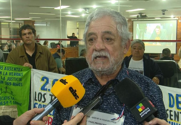 Hombre canoso con micrófono y camisa oscura, rodeado de un grupo atento en un auditorio con carteles de justicia.
