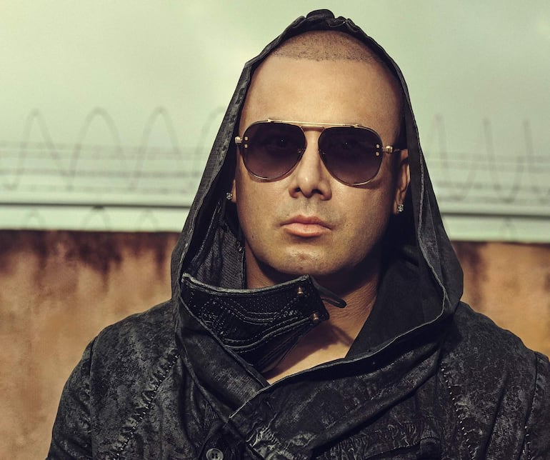 Wisin lleva gafas de sol y un abrigo elegante, posando con una actitud tranquila en el evento.