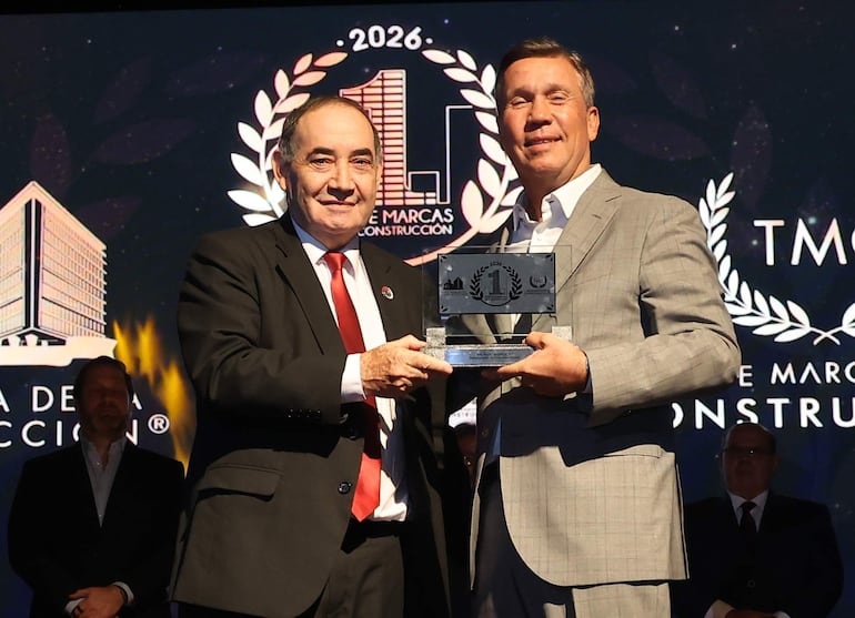 Hombre en traje negro con corbata roja entrega un trofeo a otro en traje claro, fondo decorado con alusiones a la construcción.