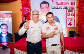 Osvaldo Sánchez y Rigoberto Chamorro sonríen con gesto de victoria, en salón decorado con globos rojos y mantel rojo y blanco.