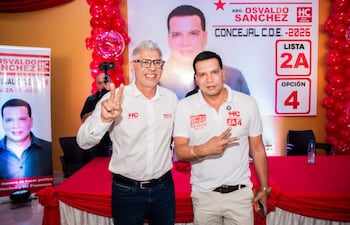 Osvaldo Sánchez y Rigoberto Chamorro sonríen con gesto de victoria, en salón decorado con globos rojos y mantel rojo y blanco.