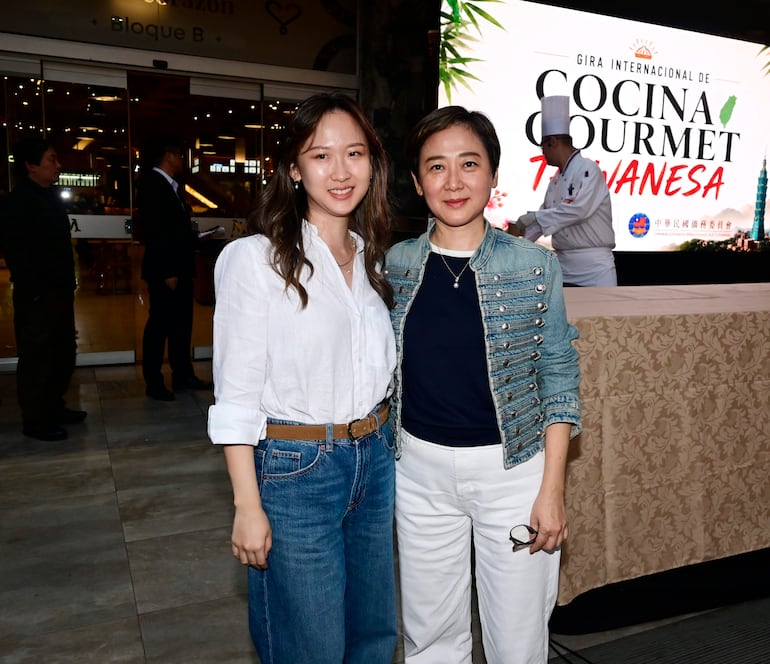 Dos mujeres sonrientes, una en blusa blanca y otra en chaqueta de mezclilla, posan en un evento culinario con un chef al fondo.
