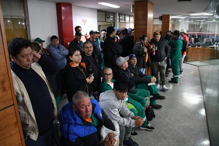 Varias personas, entre ellas Lorenzo Barreto, en tonos oscuros y verdes, esperan en una sala de espera con expresiones tranquilas.