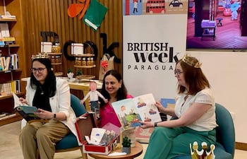 Danielle Dunne con vestimenta clara y corona, sostiene un libro ilustrado, mientras un niño le observa atento desde un salón decorado.