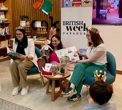 Danielle Dunne con vestimenta clara y corona, sostiene un libro ilustrado, mientras un niño le observa atento desde un salón decorado.