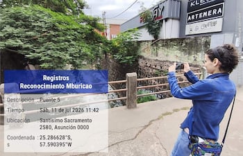 Persona de perfil con chaqueta de mezclilla, tomando fotos con su móvil frente a carteles publicitarios en un entorno urbano.