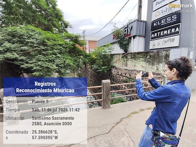 Persona de perfil con chaqueta de mezclilla, tomando fotos con su móvil frente a carteles publicitarios en un entorno urbano.