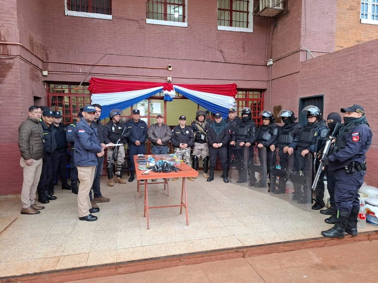 Agentes de seguridad en uniformes tácticos, algunos armados, alineados frente a un portavoz en una instalación de seguridad decorada con cinta patriótica.