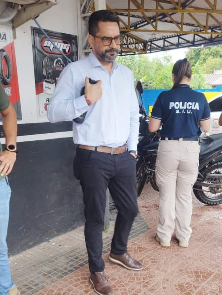 Tres personas en un puesto policial; hombre de camiseta verde observa, otro en camisa blanca sostiene un objeto y mujer con camiseta negra espera.