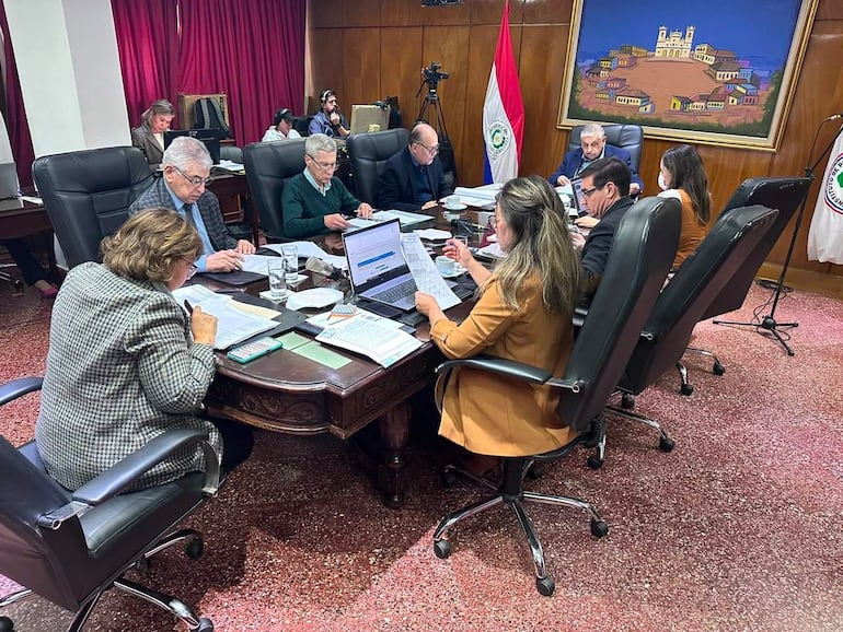 Isaías Ricardo Fretes en chaqueta oscura, rodeado de otros adultos en sala de reuniones con paredes de madera y banderas de Paraguay.