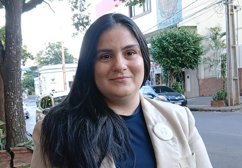 Mujer de cabello oscuro con chaqueta clara y camiseta oscura, luciendo un pin distintivo, en un ambiente urbano.