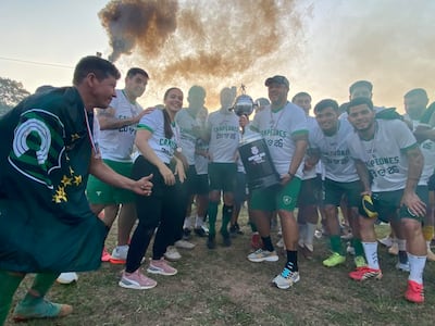Grupo de personas sonrientes con camisetas blancas que dicen 'CAMPEONES 2023', sosteniendo un trofeo en un ambiente festivo.