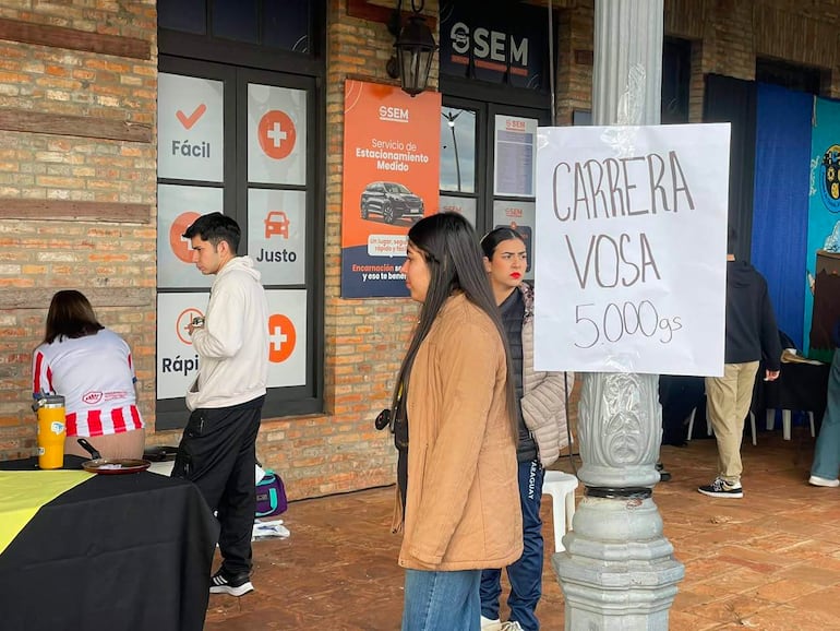 Dos mujeres, una en camiseta a rayas y otra con abrigo marrón, asisten a evento solidario con cartel de 'CARRERA VOSA' detrás.