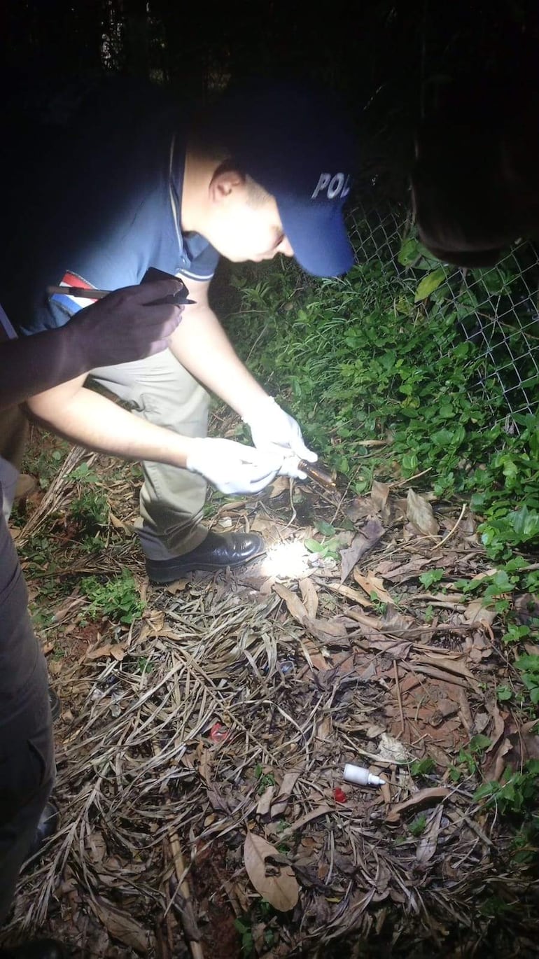 Policía en uniforme arrodillado examina el suelo con un frasco oscuro en un ambiente natural durante la noche.