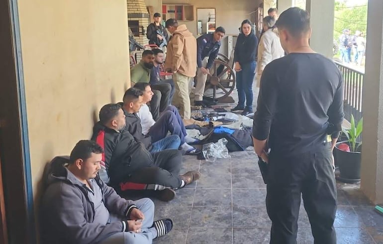 Ocho hombres sentados en el suelo, rodeados por agentes de seguridad que revisan objetos en un ambiente interior sencillo.