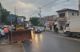 Varios trabajadores con chalecos reflectantes conversan junto a una retroexcavadora en una calle húmeda, rodeados de vehículos.