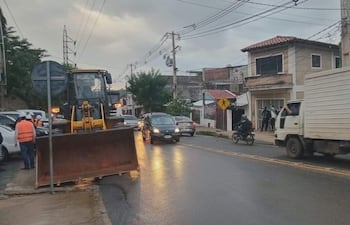 Varios trabajadores con chalecos reflectantes conversan junto a una retroexcavadora en una calle húmeda, rodeados de vehículos.