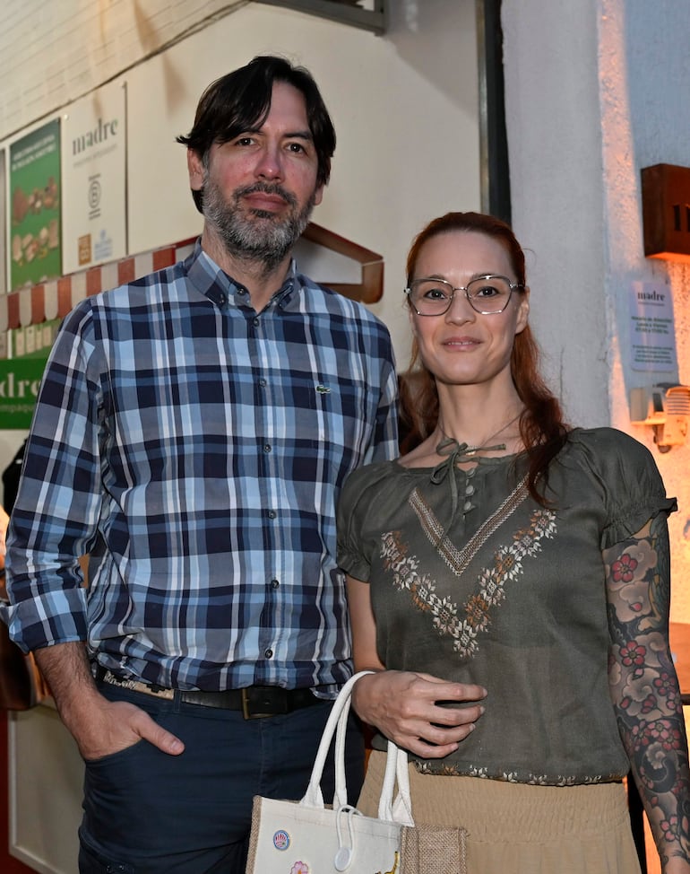 Hombre con camisa de cuadros y mujer en blusa verde y falda clara, sosteniendo un bolso, en un ambiente iluminado.