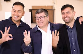 Tres hombres sonrientes en interior, mostrando dedos manchados de tinta, celebran victoria electoral con gestos de triunfo.
