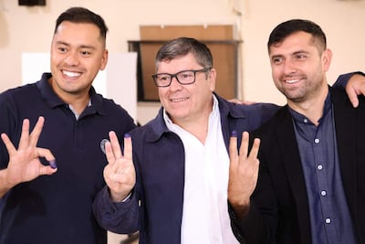 Tres hombres sonrientes en interior, mostrando dedos manchados de tinta, celebran victoria electoral con gestos de triunfo.