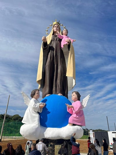 Estatua de la Virgen María rodeada de personas en oración, cielo nublado y vegetación al fondo.