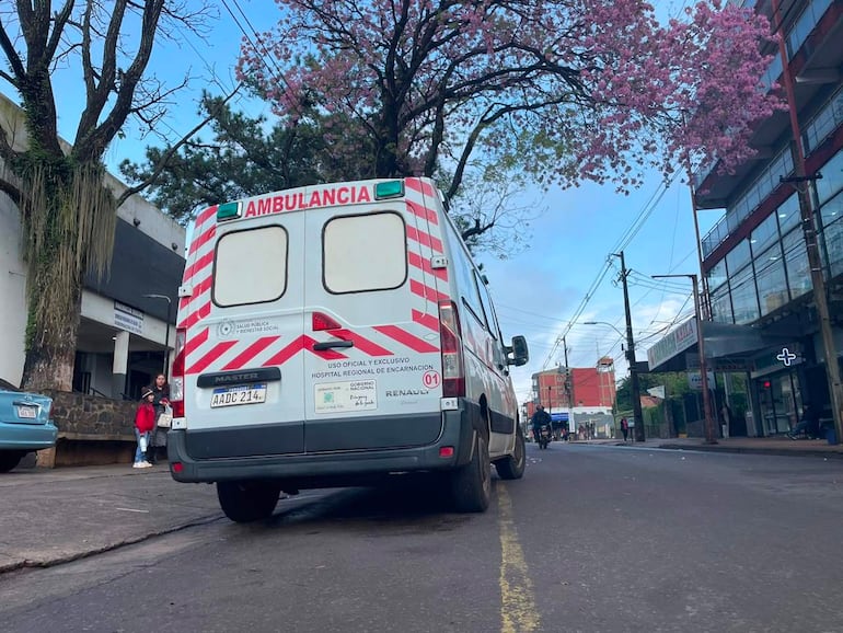 Ambulancia con inscripciones de salud pública, en una calle pavimentada con un árbol en flor al fondo.